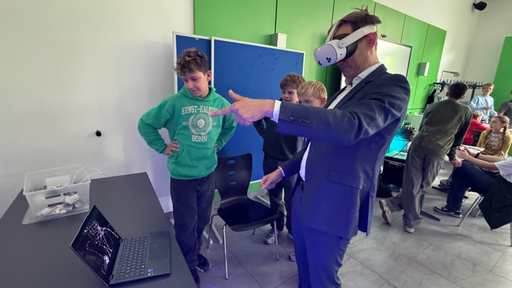 Der Bundestagsabgeordnete Hendrik Streeck steht mit einer VR-Brille auf dem Kopf in einem Klassensaal mit mehreren Schülern des Gymnasiums