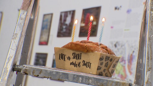 Ein Kuchen mit drei leuchtenden Kerzen steht auf einer Leiter, im Hintergrund hängen Bilder