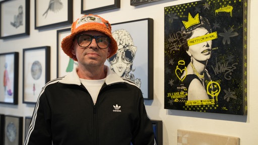 Ein Mann mit orangener Mütze steht vor einem seiner Kunstwerke