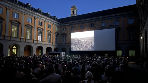 Viele Menschen sitzen in einem Innenhof und schauen auf eine große Leinwand.