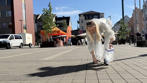 Eine Frau in einem weißen Anzug wie Tatortreiniger liest Zigaretten vom Boden auf