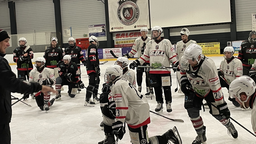 Mehrere männliche Eishockey-Spieler stehen in Schutzausrüstung auf der Eisbahn.