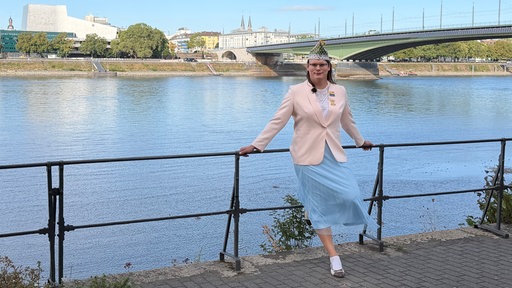 Frau in rosa Jackett und mit Karnevalsschiffchen auf dem Kopf lehnt an Geländer am Rhein