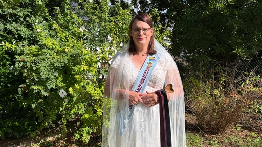 Frau in Brautkleid, Schleier und blauer Schärpe hält einen Kochlöffel mit drangebundenen Krawatten 