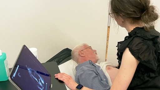 Ein Mann liegt mit entblößter Brust auf einer Liege, darüber ist eine Ärztin in dunkler Kleidung gebeugt und berührt mit der linken Hand einen Ultraschall-Monitor.