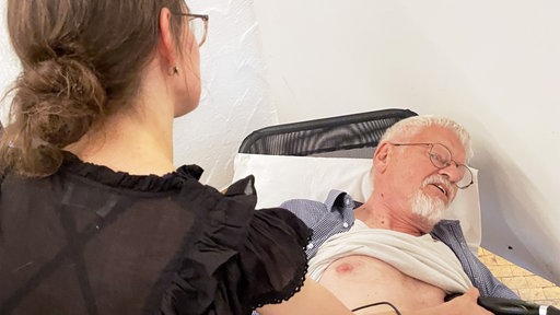 Ein Mann liegt mit entblößter Brust auf einer Liege, links darüber ist eine Ärztin in dunkler Kleidung gebeugt und hält mit in der rechten Hand ein schwarzes Ultraschallgerät auf die Brust des Mannes.