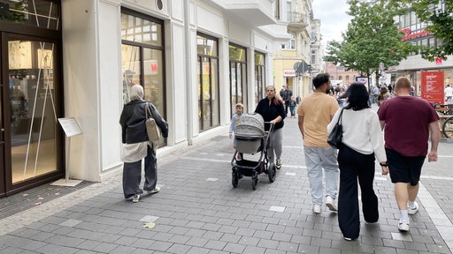 Eine gepflasterte Fußgängerzone in Bonn, Passanten laufen an einem weißen Gebäude mit hohen Schaufenstern vorbei.