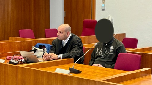 In einem Gerichtssaal sitzen ein Anwalt und ein Angeklagter. Das Gesicht des Angeklagten ist unkenntlich gemacht.