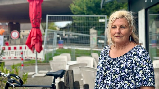 Eine Frau mit blonden Haaren schaut rechts an der Kamera vorbei. Im Hintergrund sind Bauzäune und Absperrungen.