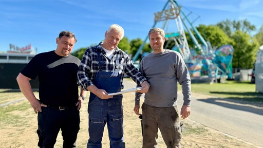 Drei Männer stehen vor einer Schiffschaukel in der Rheinaue.