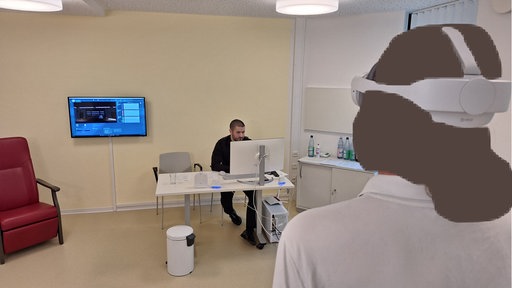 Ein Patient trägt eine VR-Brille, ihm gegenüber ist ein Mann an einem Schreibtisch mit Rechner, an der Wand sieht man einen Bildschirm