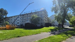Bauarbeiter auf einem Gerüst vor einem Mehrfamilienhaus. Im Vordergrund stehen ein Kran und ein gelber Schuttcontainer.
