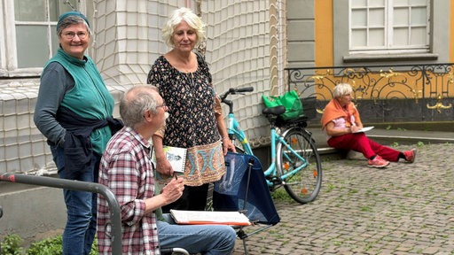 Zwei Frauen und ein Mann stehen vor einem gelben Gebäude und halten Zeichenblöcke.