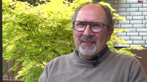 Mann mit Brille und grau-weißem Bart, Mitglied der Bürgerinitiative, trägt einen beigen Pullover, im Hintergrund gelb-grüne Sträucher und eine weiße Mauer