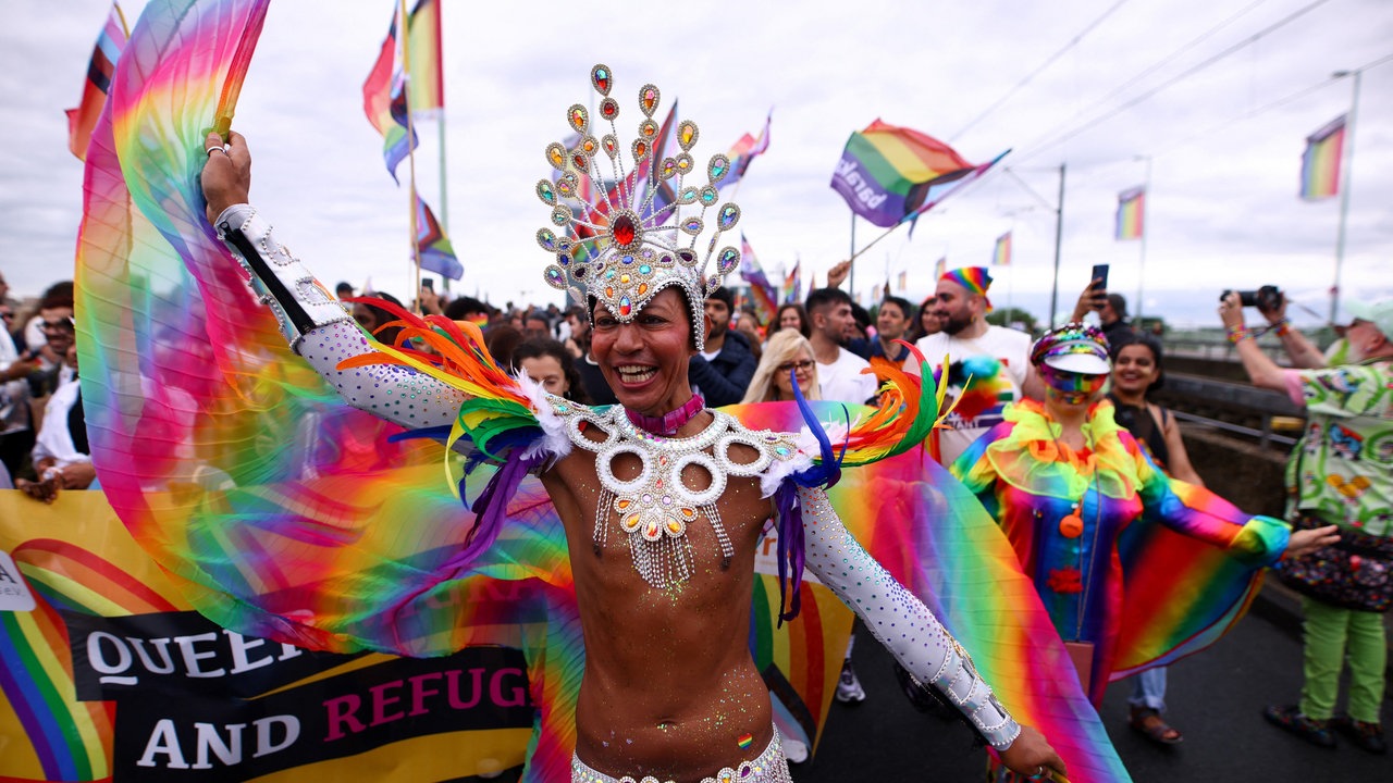 Eine Drag-Queen mit Regenbogen-Flügeln, viel Glitzer und freiem Oberkörper auf der Deutzer Brücke, im Hintergrund Menschenmengen