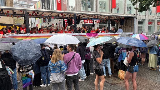 Menschen mit Regenschirmen stehen am Rand der CSD-Parade in Köln