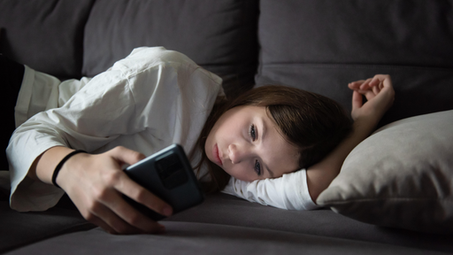 Ein trauriges Teenager-Mädchen liegt mit einem Smartphone in der Dunkelheit (Symbolbild)