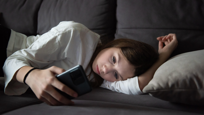 Ein trauriges Teenager-Mädchen liegt mit einem Smartphone in der Dunkelheit (Symbolbild)