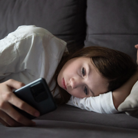Ein trauriges Teenager-Mädchen liegt mit einem Smartphone in der Dunkelheit (Symbolbild)