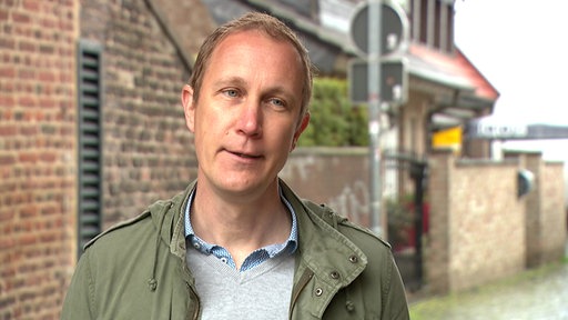 "Daniel Zimmermann, ehemaliger Bürgermeister von Monheim am Rhein, lächelt im Freien; er trägt ein gemustertes Hemd unter einer olivgrünen Jacke, im Hintergrund eine unscharfe Straßenszene.