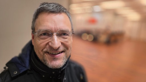 EIn Mann mit durchsichtiger Brille, Bart und grauem Haar in einem Saaleingang