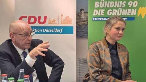 Ein Mann und eine Frau an einem Tisch, dahinter die jeweiligen Parteilogos