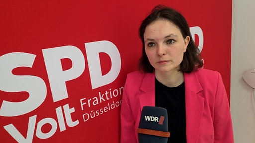 Eine Frau in schwarzem T-Shirt und rosa Blazer vor einem roten Parteilogo