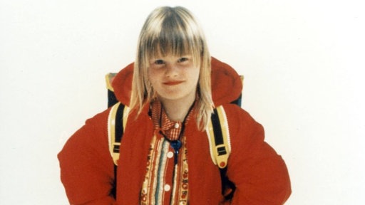 Ein Mädchen mit blondem, schulterlangem Haar, das eine rote Jacke, einen Rucksack und ein gemustertes Oberteil trägt und vor hellem Hintergrund in die Kamera lächelt.