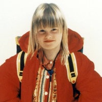 Ein Mädchen mit blondem, schulterlangem Haar, das eine rote Jacke, einen Rucksack und ein gemustertes Oberteil trägt und vor hellem Hintergrund in die Kamera lächelt.