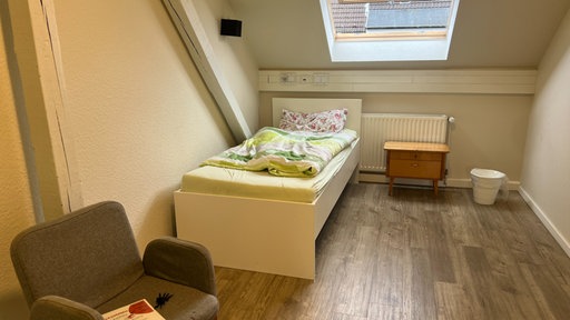 Ein mit bunter Bettwäsche bezogenes Bett, außerdem ein Nachttisch und ein Sessel.