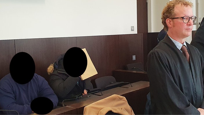 Zwei angeklagte Männer in dunkler Kleidung sitzen hinter einem Holztisch im Gerichtssaal, ihre Gesichter sind unkenntlich gemacht. Rechts im Bild steht der Verteidiger, ein Mann in dunkler Robe.
