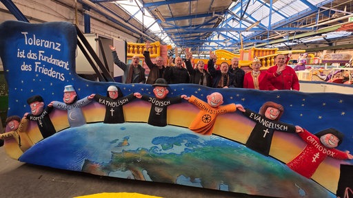 Ein Karnevalswagen in einer großen Halle mit winkenden Menschen drauf.