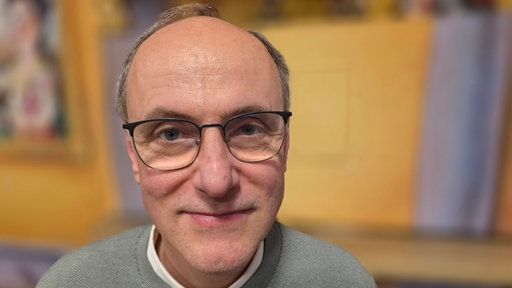 Ein Mann schaut in die Kamera, er trägt eine Brille und einen dunkelgrünen Pullover