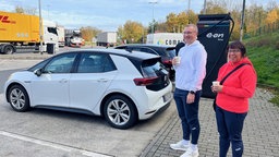 Ein Mann und eine Frau stehen an einer e-Auto Ladestation, warten und trinken einen Kaffee
