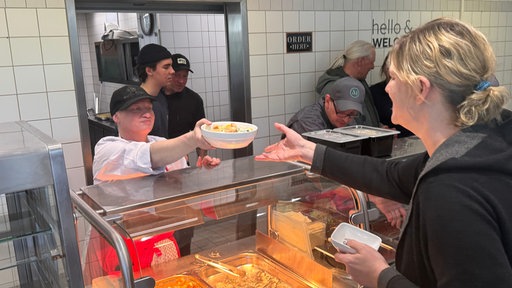 Eine Frau reicht in der Kantine des NRW-Sozialministeriums eine Schüssel mit Essen über die Theke. Eine andere Frau nimmt sie entgegen.