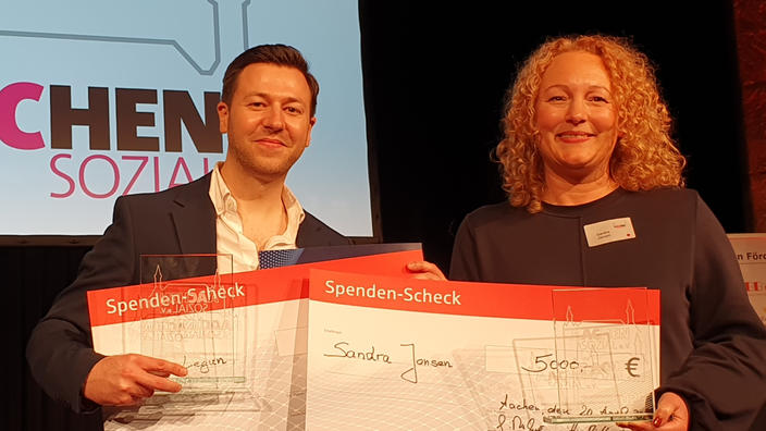 Ein Mann mit kurzen, schwarzen Haaren und eine Frau mit roten, lockigen Haaren halten jeweils einen großen Spenden-Scheck mit der Aufschrift "5000 Euro" in den Händen.