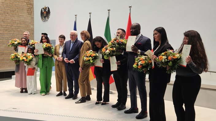 Die neu-eingebürgerten Personen stehen u.a. mit Bundespräsident Steinmeier in einer Reihe und halten ihre Urkunden sowie Blumensträuße in den Händen.