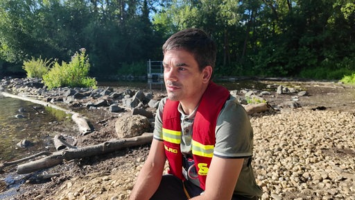 Ein Mann in roter DLRG-Weste hockt am Ufer eines Gewässers.