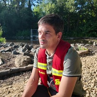 Ein Mann in roter DLRG-Weste hockt am Ufer eines Gewässers.