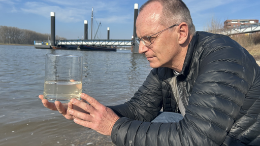 Dirk Jansen hockt neben einer Stelle am Rhein und hält in der Hand ein Glas mit Wasser aus dem Fluss