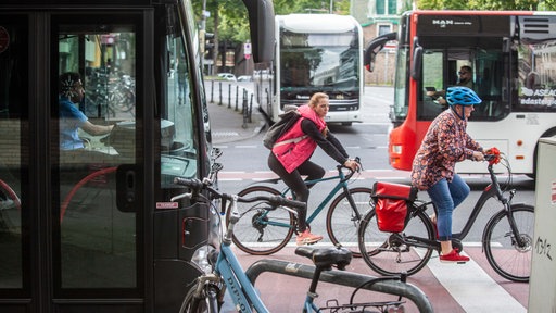 Es sind zwei Radfahrer zu sehen, die auf einem Radweg neben mehreren Linienbussen unterwegs sind.