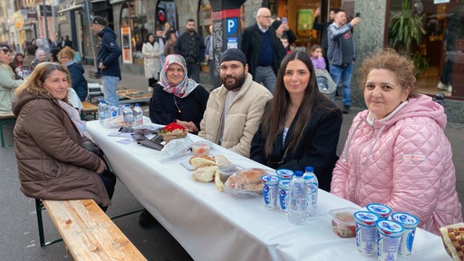 Fünf Menschen sitzen an einem Tisch auf einer Straße. Auf dem Tisch liegen verschiedene Speisen