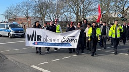 Ein Demozug mit teils in Warnwesten bekleideten Ford-Mitarbeitern zieht an einem Polizeiwagen vorbei. Vorne haben sie ein Banner mit der Aufschrift "Wir bleiben Ford"