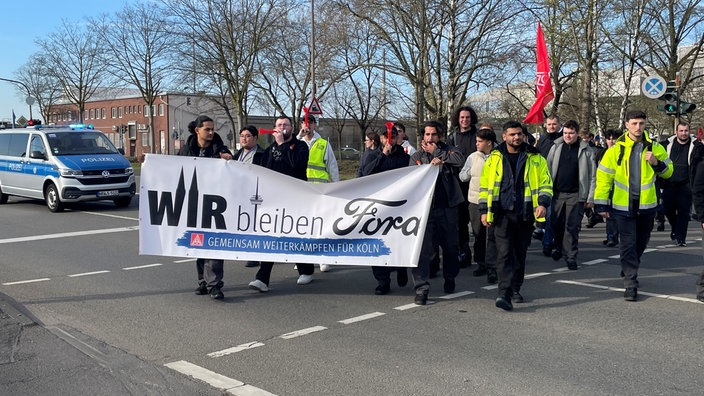 Ein Demozug mit teils in Warnwesten bekleideten Ford-Mitarbeitern zieht an einem Polizeiwagen vorbei. Vorne haben sie ein Banner mit der Aufschrift "Wir bleiben Ford"