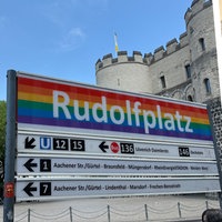 Die linke Bildhälfte zeigt das Haltestellen-Schild "Rudolfplatz" in regenbogenfarben. Rechts steht Jenny.