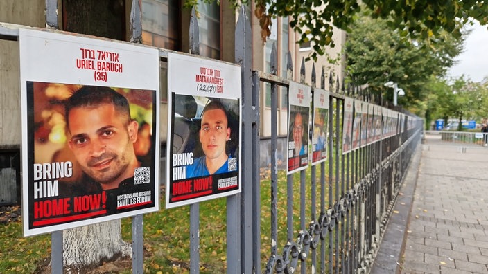 Zu sehen sind an einem Zaun hängende Plakate, auf denen jeweils die Geiseln abgebildet sind.