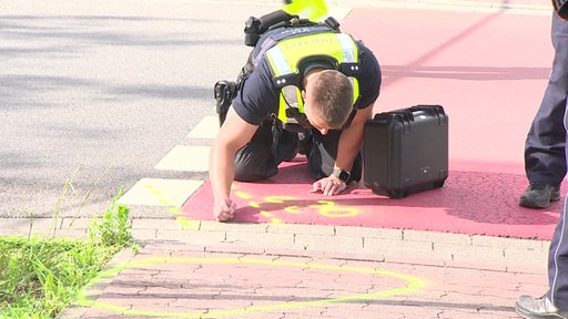 Ein Polizist kniet auf dem Radweg und beugt sich über neongelbe Markierungen