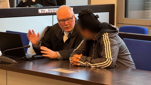 Auf dem Foto ist die Angeklagte: eine Frau mit Adidas-Jacke, schwarzen Haaren und weiß lackierten Fingernägeln. Daneben ihr Verteidiger.