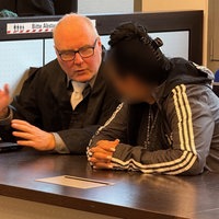 Auf dem Foto ist die Angeklagte: eine Frau mit Adidas-Jacke, schwarzen Haaren und weiß lackierten Fingernägeln. Daneben ihr Verteidiger.