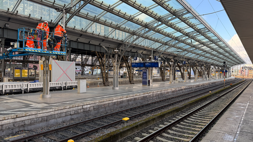 Arbeiter in orangenen Anzügen stehen auf einer Hebebühne neben den Gleisen am Hauptbahnhof.